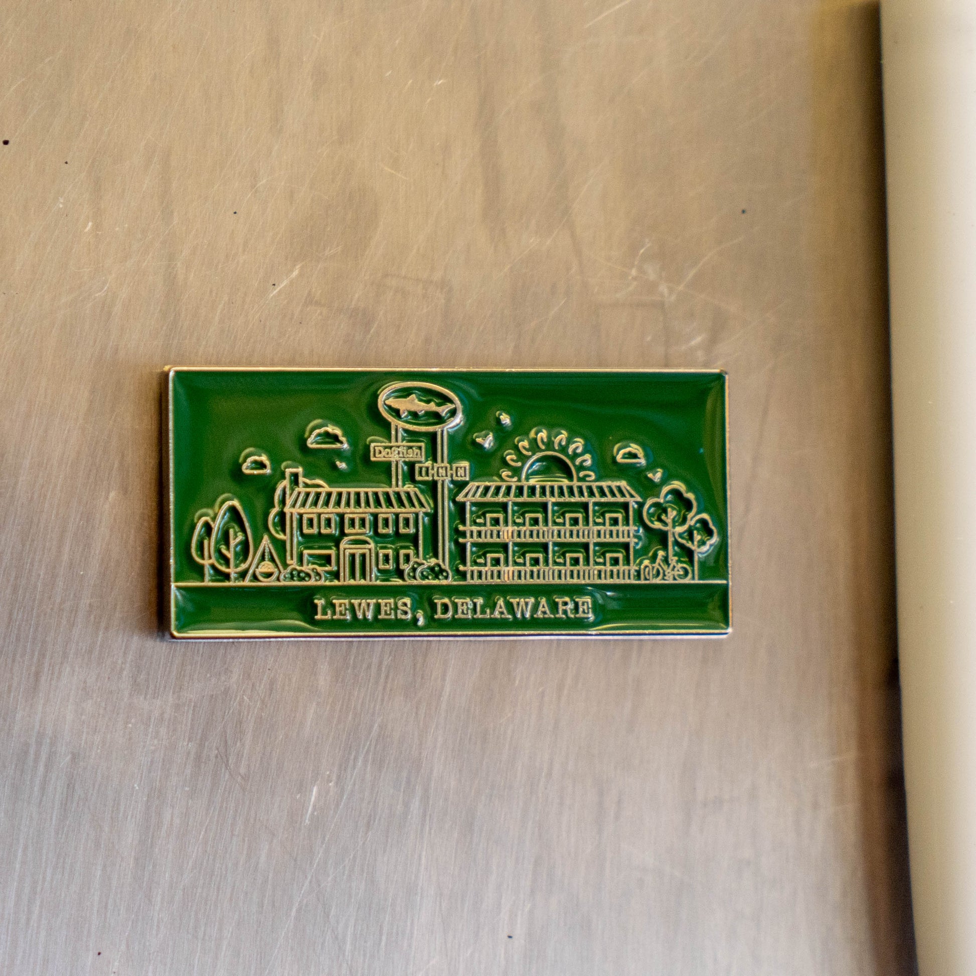 The Enamel Inn Magnet by Arrasmith Promotions LLC is a green rectangular magnet featuring buildings and trees, inspired by the charm of Lewes, Delaware.