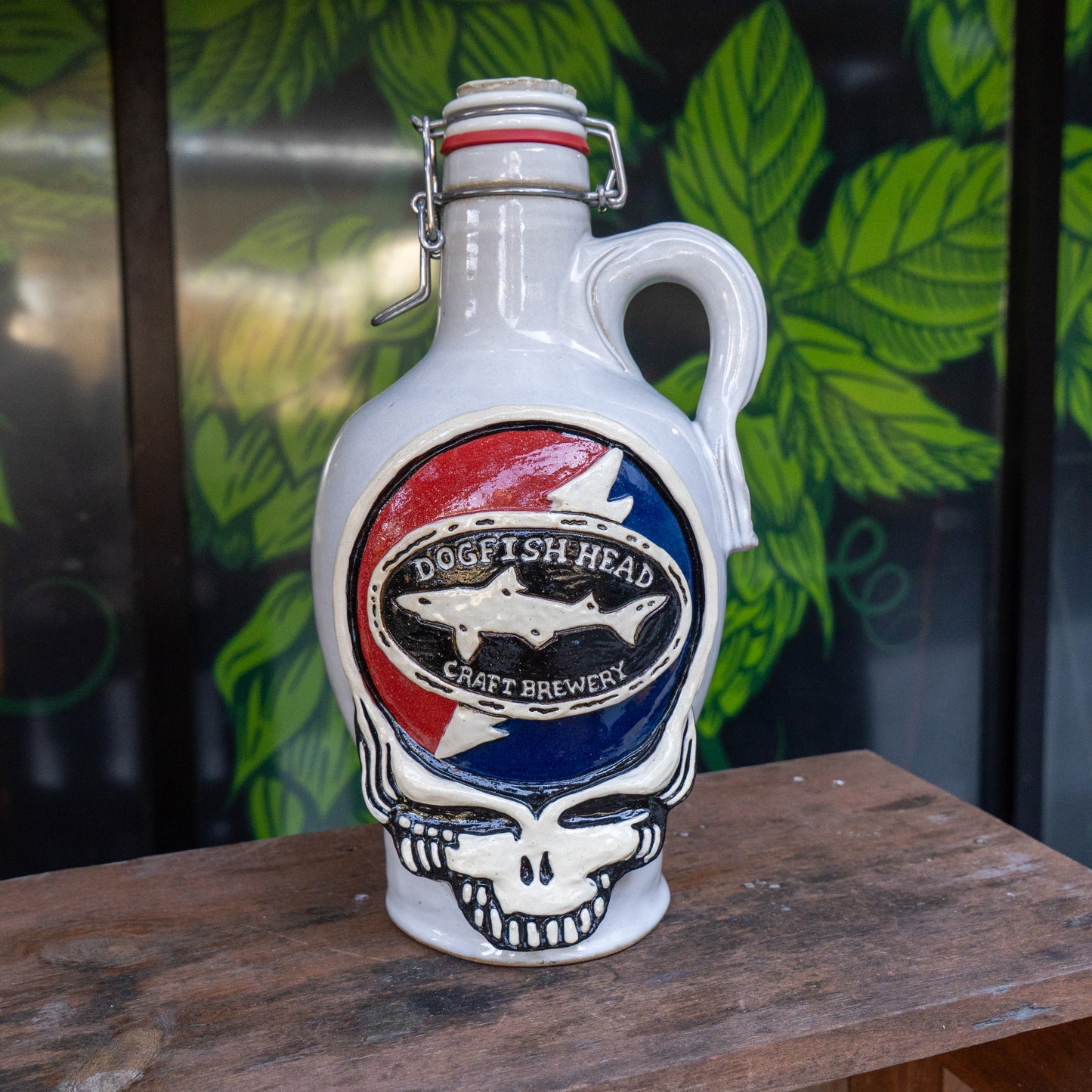 The Uber Growler by Romanick Pottery, featuring a skull design, rests on a wooden surface with green leaves painted on a dark backdrop behind it.