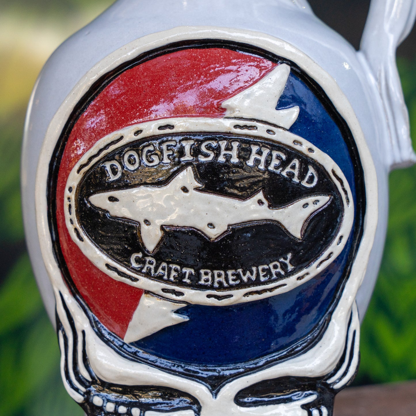 Close-up of the Uber Growler by Romanick Pottery, featuring an oval logo with a shark silhouette and the words Dogfish Head Craft Brewery on a red and blue background, framed by a white skeleton-like border.