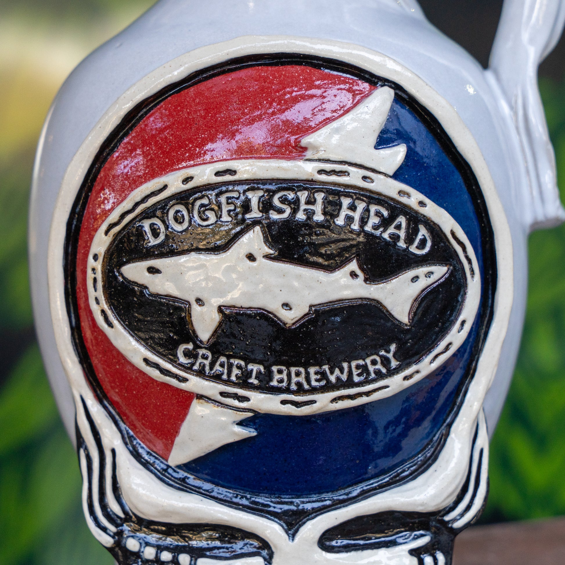 Close-up of the Uber Growler by Romanick Pottery, featuring an oval logo with a shark silhouette and the words Dogfish Head Craft Brewery on a red and blue background, framed by a white skeleton-like border.