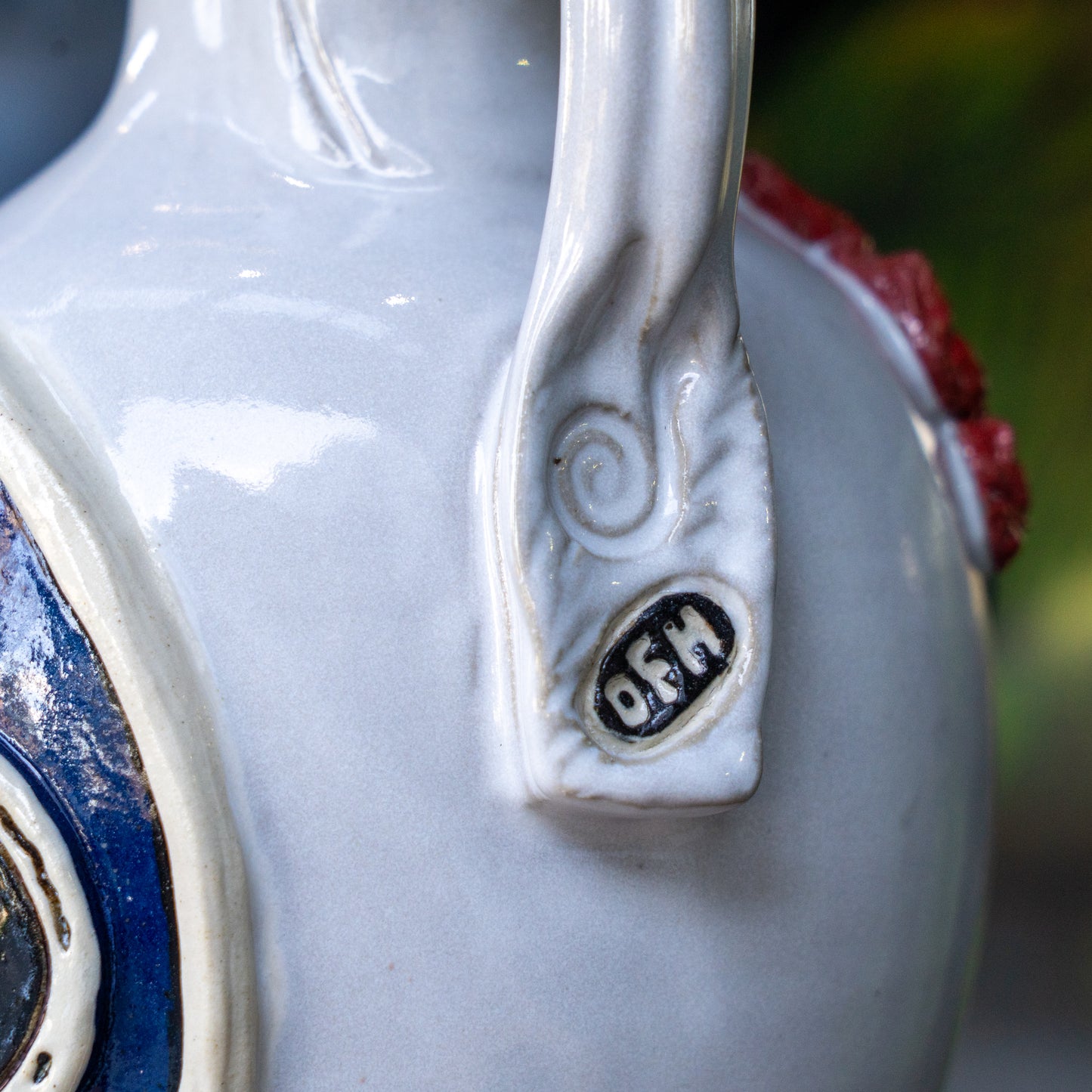 Close-up of the Uber Growler by Romanick Pottery, featuring a handmade spiral handle with "OFM" engraved in black on a white oval. The glossy light gray ceramic has blue and red decorative accents.