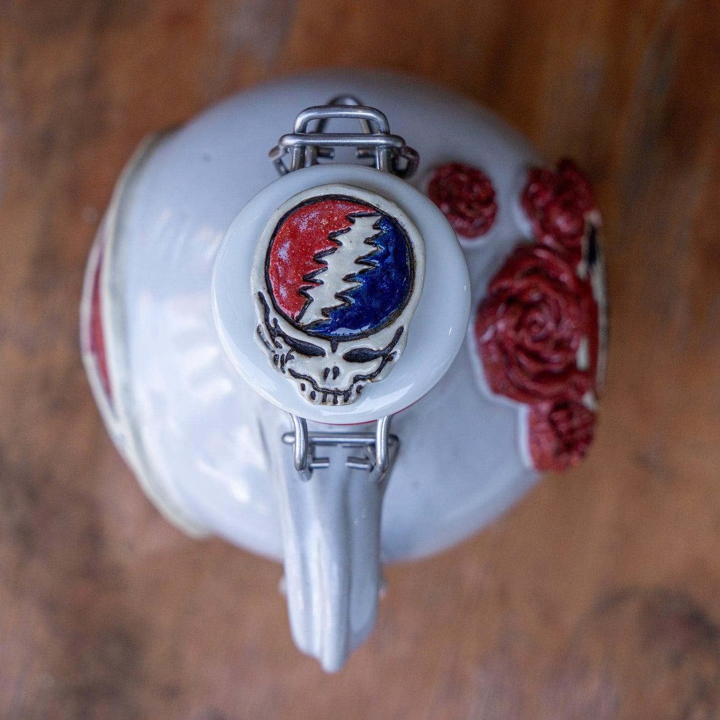 The Uber Growler by Romanick Pottery is a ceramic growler with a metal clasp, decorated with a skull, red and blue lightning bolt, and red roses, photographed from above on a wooden surface.