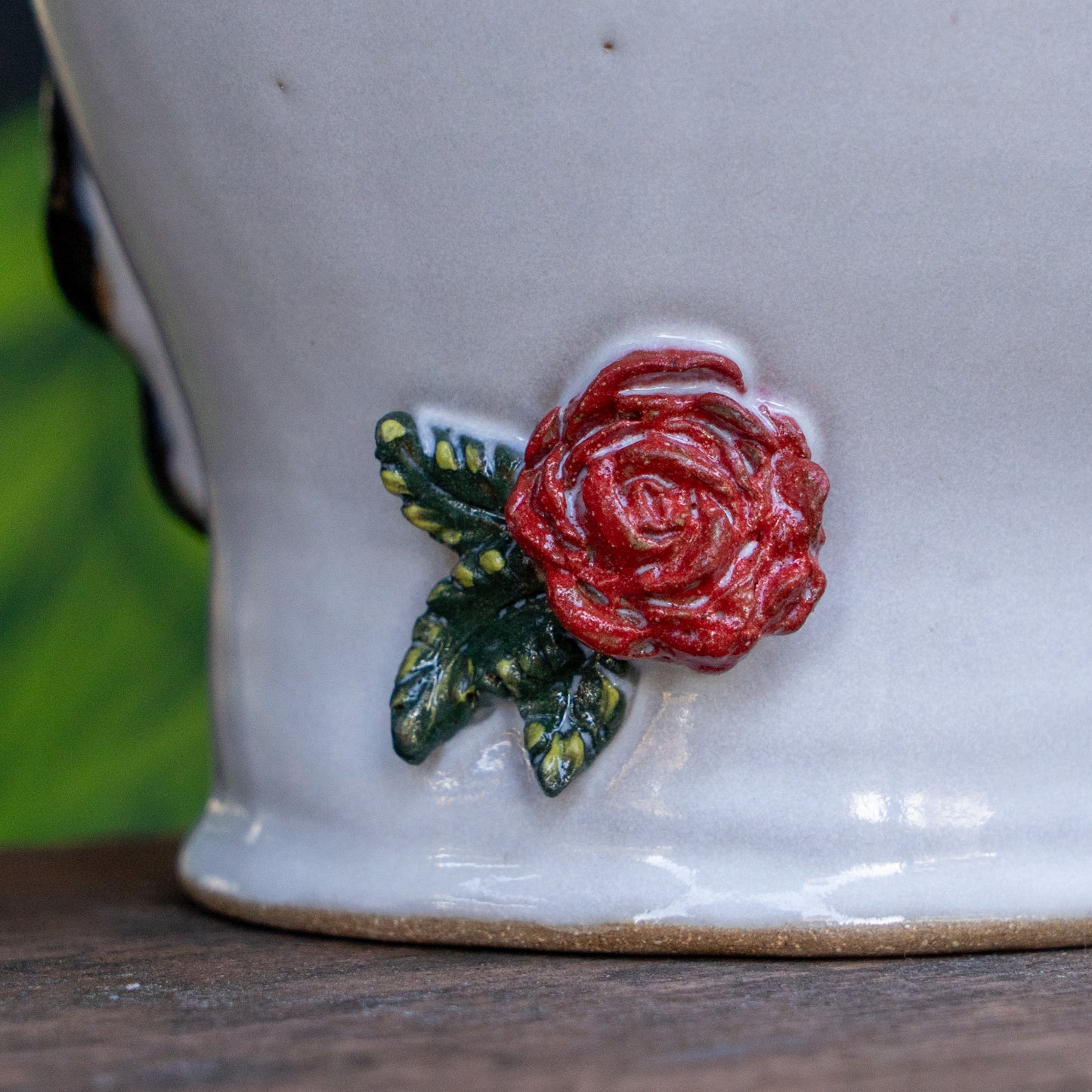 A close-up of the Uber Growler by Romanick Pottery—a white handmade pot with a raised red rose and green leaves—sits on a wooden surface, blurred greenery in the background.