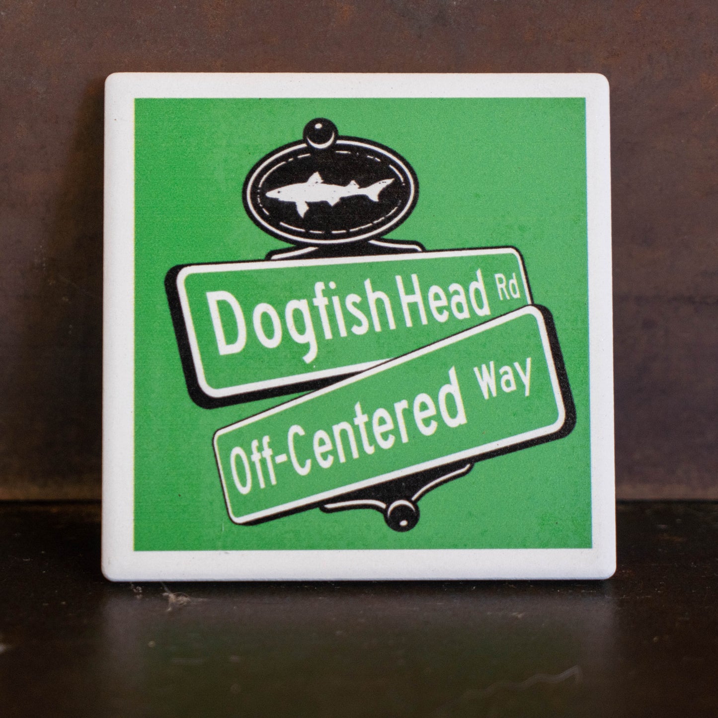A square ceramic tile coaster featuring a green and white street sign design with the text 'Dogfish Head Rd' and 'Off-Centered Way' on angled green street sign panels, with a black fish emblem at the top. The tile is photographed standing upright on a dark brown wooden surface.