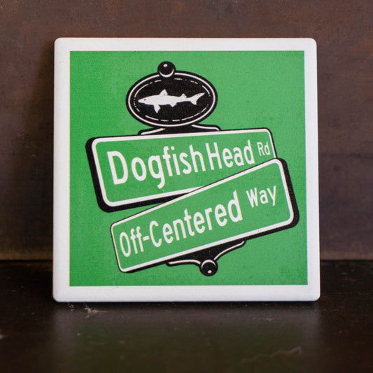 A square ceramic tile coaster featuring a green and white street sign design with the text 'Dogfish Head Rd' and 'Off-Centered Way' on angled green street sign panels, with a black fish emblem at the top. The tile is photographed standing upright on a dark brown wooden surface.
