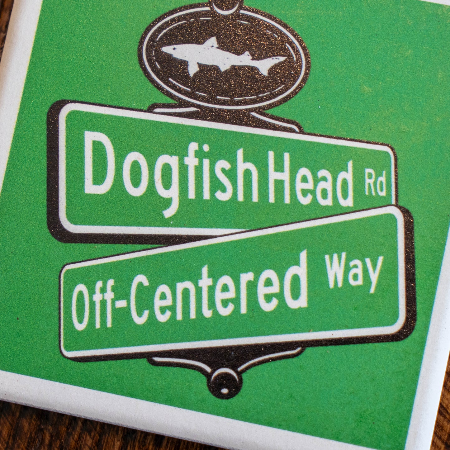 A Green Street Sign Ceramic Coaster by Paint the Town, Inc. features an illustrated street sign for Dogfish Head Rd and Off-Centered Way, topped with a shark-shaped sign, and is displayed on a wooden surface.