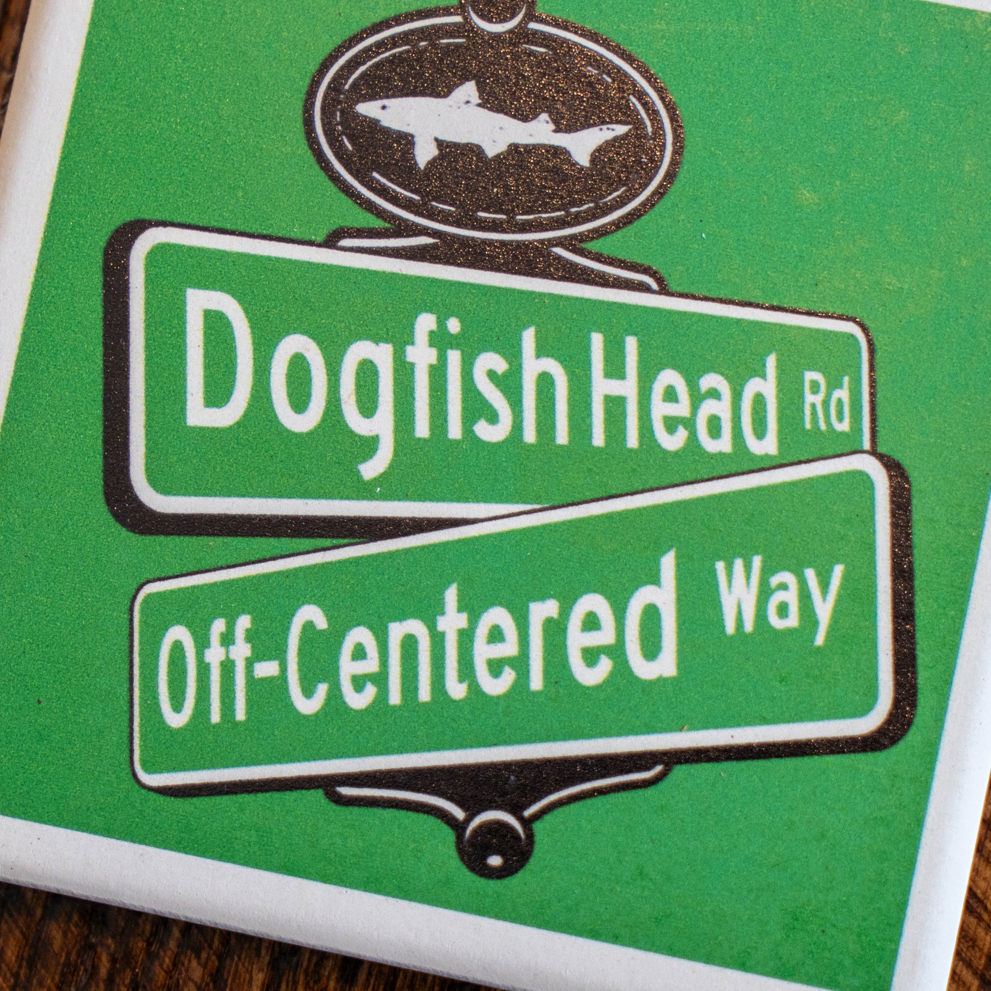 A Green Street Sign Ceramic Coaster by Paint the Town, Inc. features an illustrated street sign for Dogfish Head Rd and Off-Centered Way, topped with a shark-shaped sign, and is displayed on a wooden surface.