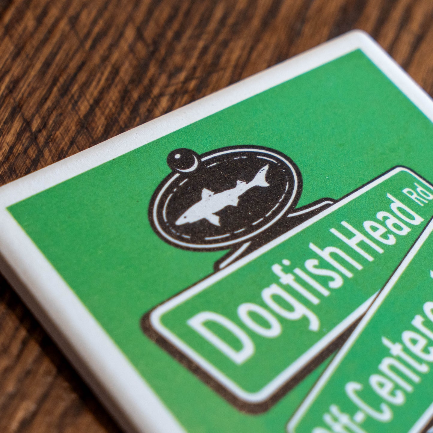 A close-up of a square coaster featuring a green and white Dogfish Head restaurant design, resting on a dark wood surface.