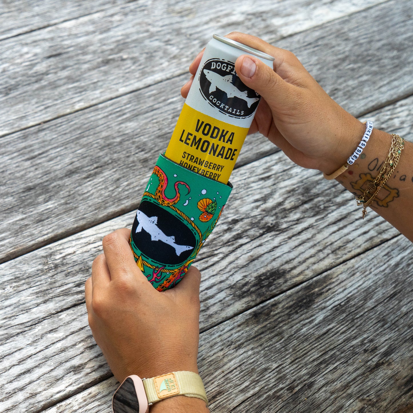 A person holds a Dogfish Head Vodka Lemonade can, wrapped in a Green Underwater Slim Can Cooler from Grandstand Glassware & Apparel, over a weathered wooden table. The can’s label highlights strawberry and honeyberry flavors, showcasing Dogfish Head’s unique approach to canned cocktails.