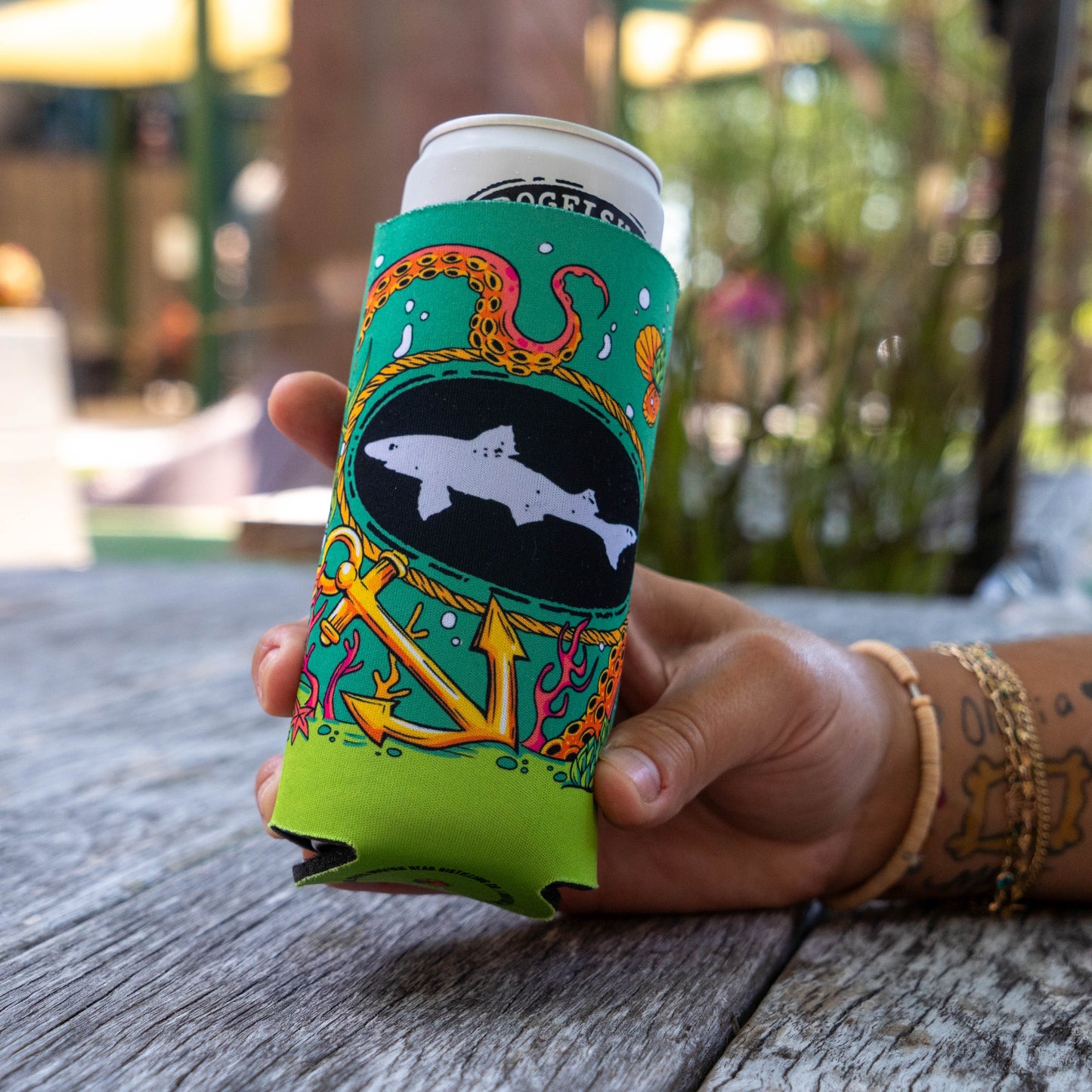 A hand holding a can cooler sleeve on a can, set on a wooden table with a blurred outdoor background. The sleeve features a green and lime green design with an anchor, a shark, and a snake.
