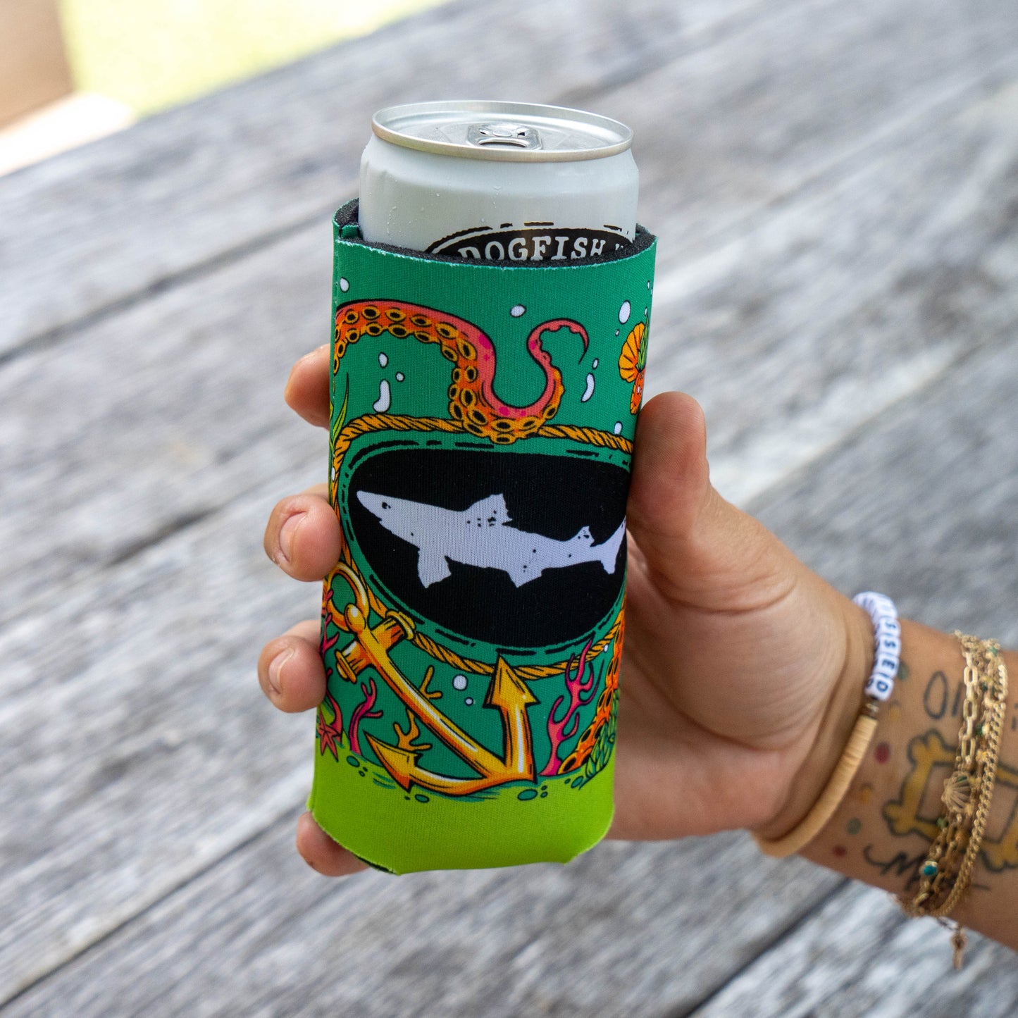 A hand holding a silver can inside a bright green neoprene can cooler with a colorful octopus and shark graphic, set against a blurred gray wooden background.