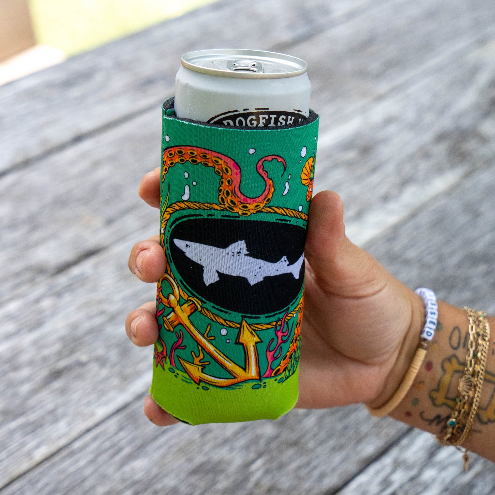 A hand holding a silver can inside a bright green neoprene can cooler with a colorful octopus and shark graphic, set against a blurred gray wooden background.