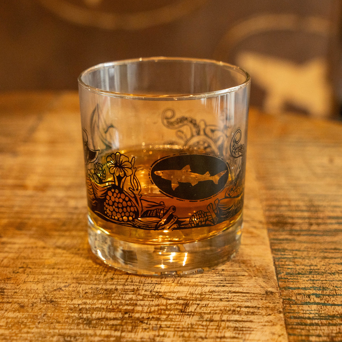 The Hull and Helm Rocks Glass by Grandstand Glassware & Apparel rests on a reflective surface, holding amber whiskey. Black octopus tentacle and sea plant illustrations decorate the glass; the background is softly blurred.