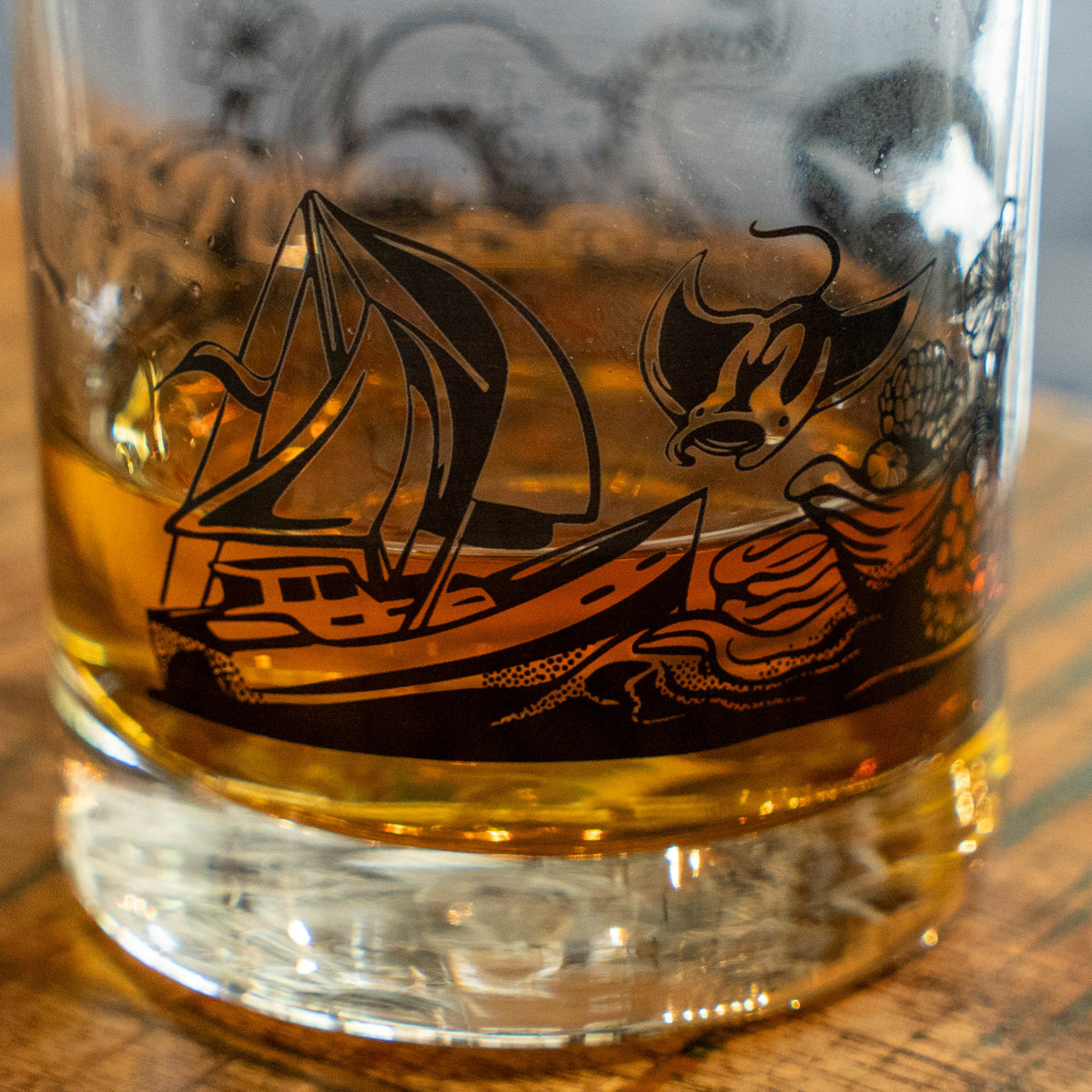The Hull and Helm Rocks Glass by Grandstand Glassware & Apparel rests on a reflective surface, holding amber whiskey. Black octopus tentacle and sea plant illustrations decorate the glass; the background is softly blurred.