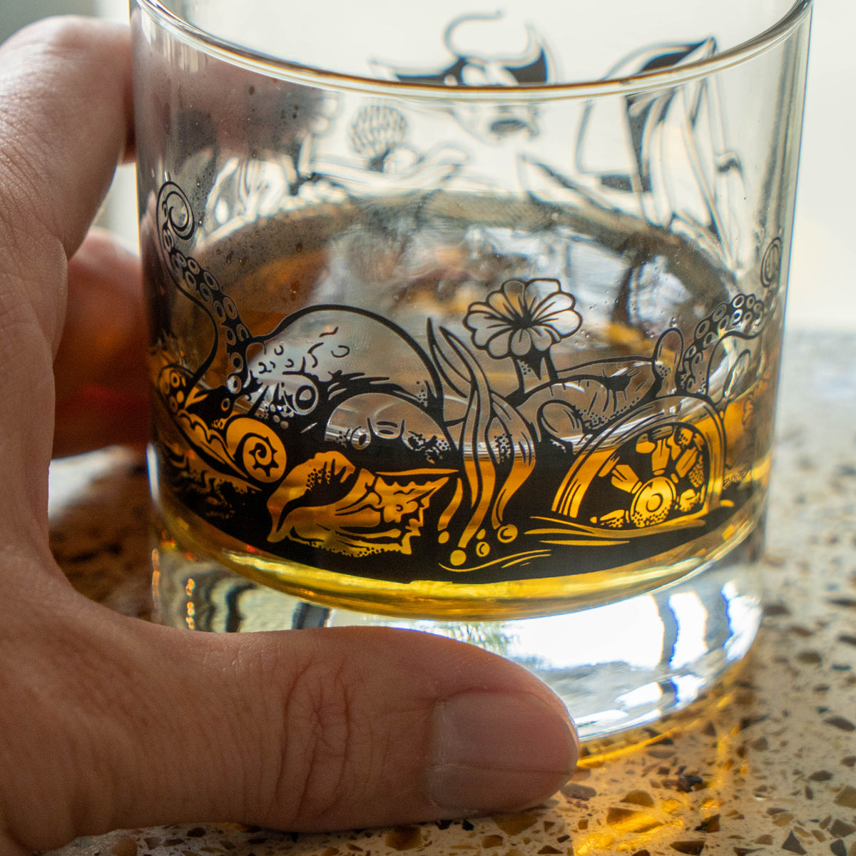 The Hull and Helm Rocks Glass by Grandstand Glassware & Apparel rests on a reflective surface, holding amber whiskey. Black octopus tentacle and sea plant illustrations decorate the glass; the background is softly blurred.