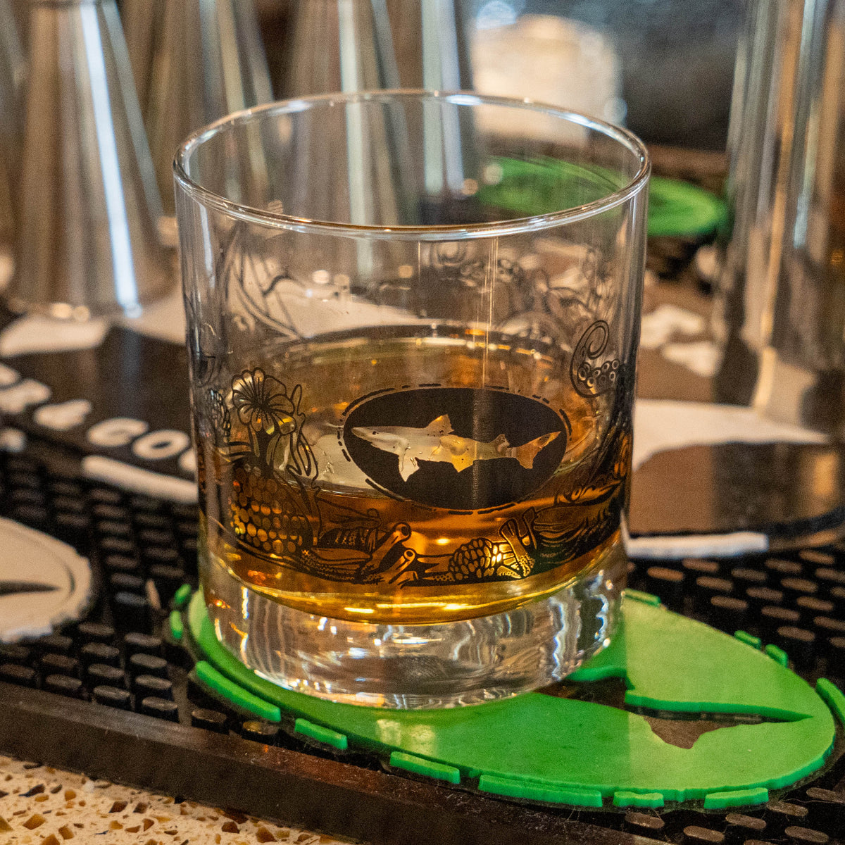 The Hull and Helm Rocks Glass by Grandstand Glassware & Apparel rests on a reflective surface, holding amber whiskey. Black octopus tentacle and sea plant illustrations decorate the glass; the background is softly blurred.