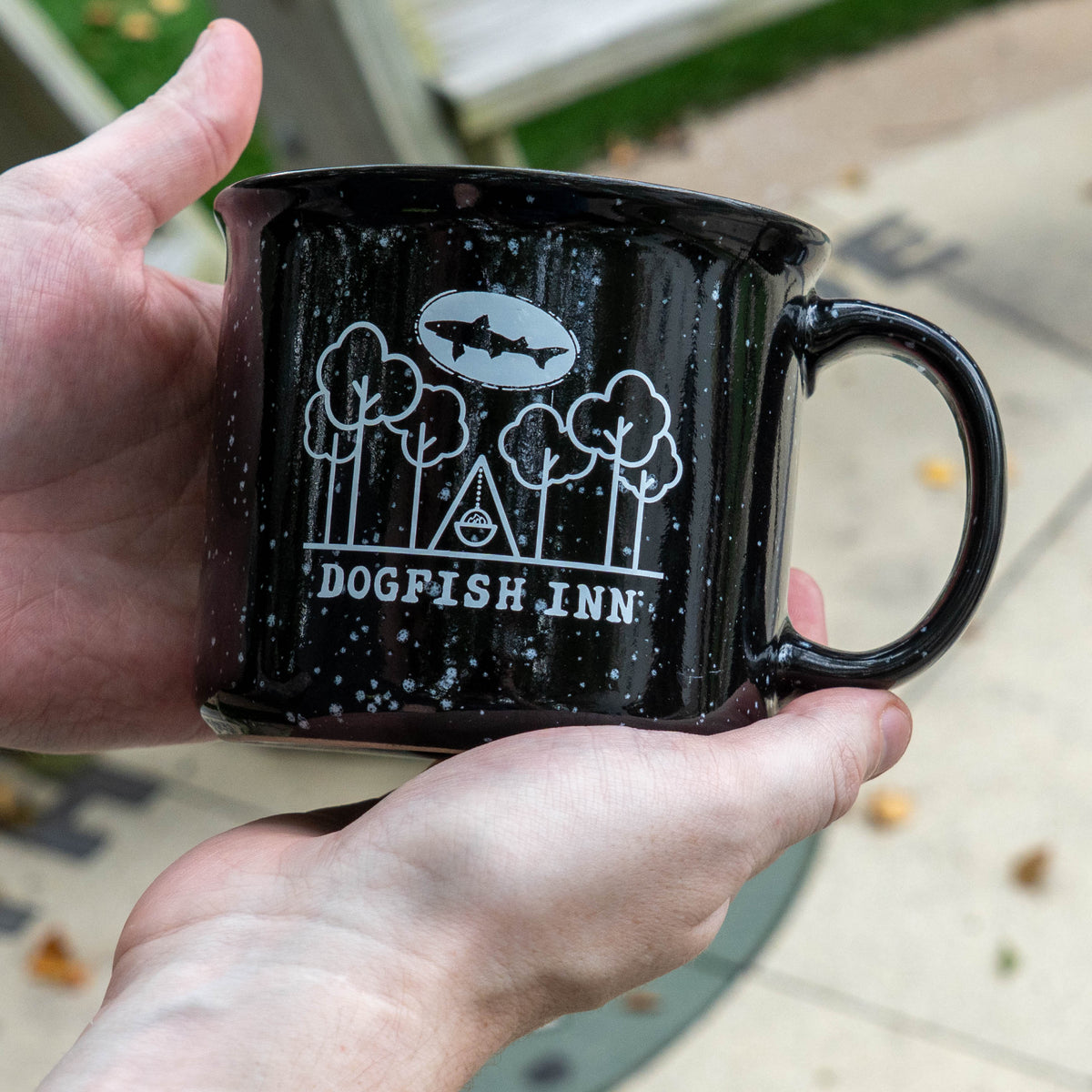 Black speckled mug being held by person with white artwork print.  Art says 'dogfish inn'