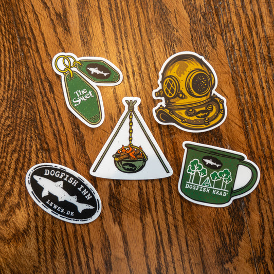 Inn Sticker Pack of 5 stickers. A black classic dogfish logo sticker, a green dogfish head camp mug sticker, a yellow-gold scuba diving mask sticker, a green keychain with two diamond shape tags (one reads "The Sweet", the other has a dogfish head logo) sticker, and a green hanging firepit bowl sticker.