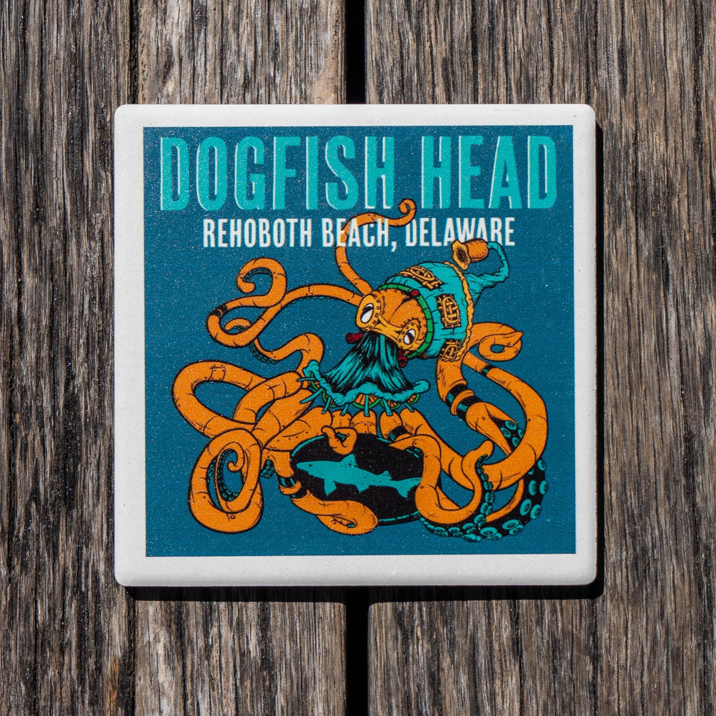 A square ceramic coaster featuring a colorful illustration of an octopus wearing a pirate hat, with the text 'DOGFSH HEAD' and 'REHOBOOTH BEACH, DELAWARE' on a teal background, shown on a dark wood surface.