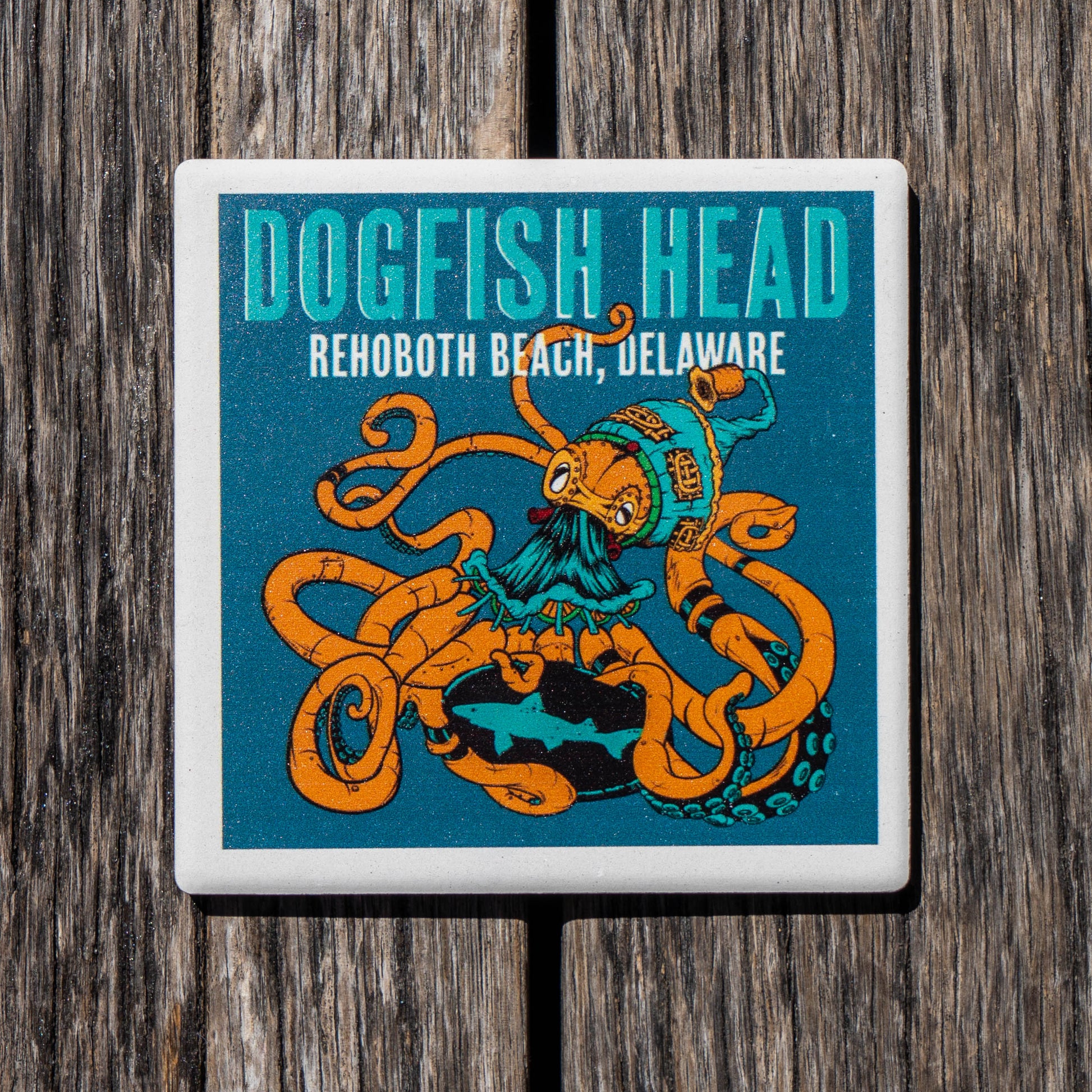 A square ceramic coaster featuring a colorful illustration of an octopus wearing a pirate hat, with the text 'DOGFSH HEAD' and 'REHOBOOTH BEACH, DELAWARE' on a teal background, shown on a dark wood surface.