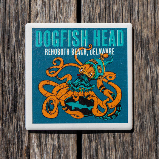 A square ceramic coaster featuring a colorful illustration of an octopus wearing a pirate hat, with the text 'DOGFSH HEAD' and 'REHOBOOTH BEACH, DELAWARE' on a teal background, shown on a dark wood surface.