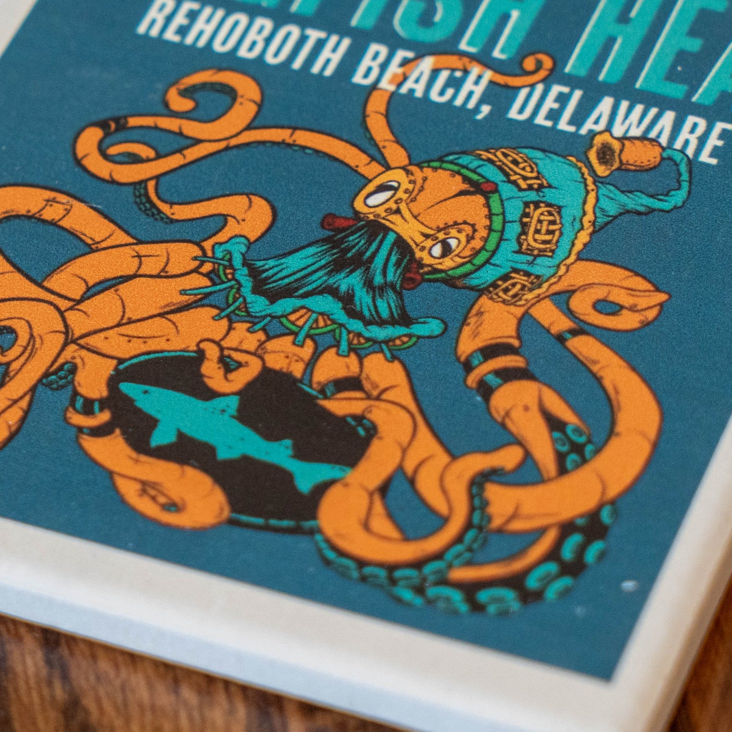 Close-up of a printed coaster or tile featuring an illustrated orange octopus wearing a teal hat and holding a black-and-teal fish on a blue background, with partial text reading 'REHOBOOTH BEACH, DELAWARE' 