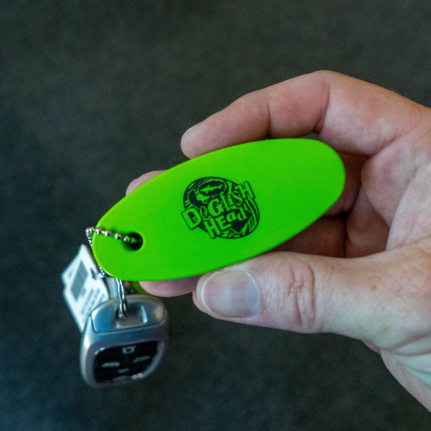 A hand holding a bright neon green keychain with a black printed 'Dogfish Head' logo, attached to a keyring with keys, against a dark gray background.
