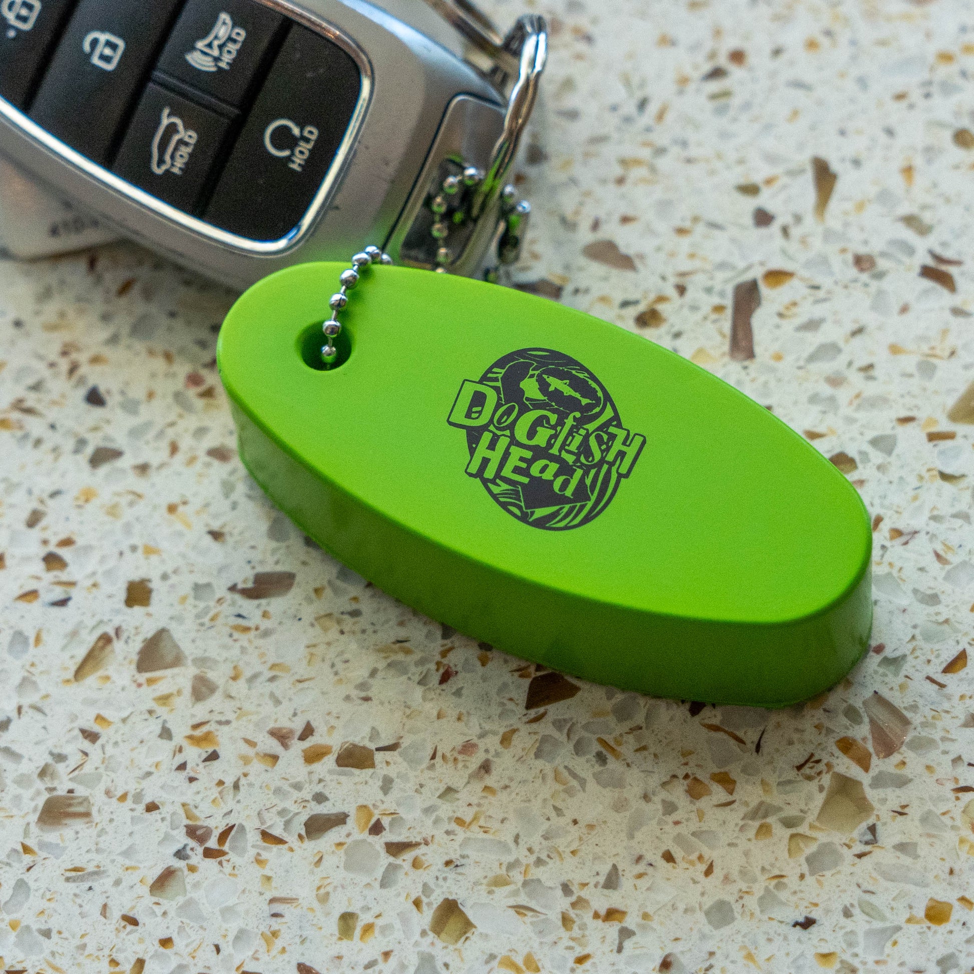 A bright lime green oval-shaped  keychain with a silver ball chain, featuring the 'Dogfish Head' logo, resting on a speckled terrazzo 