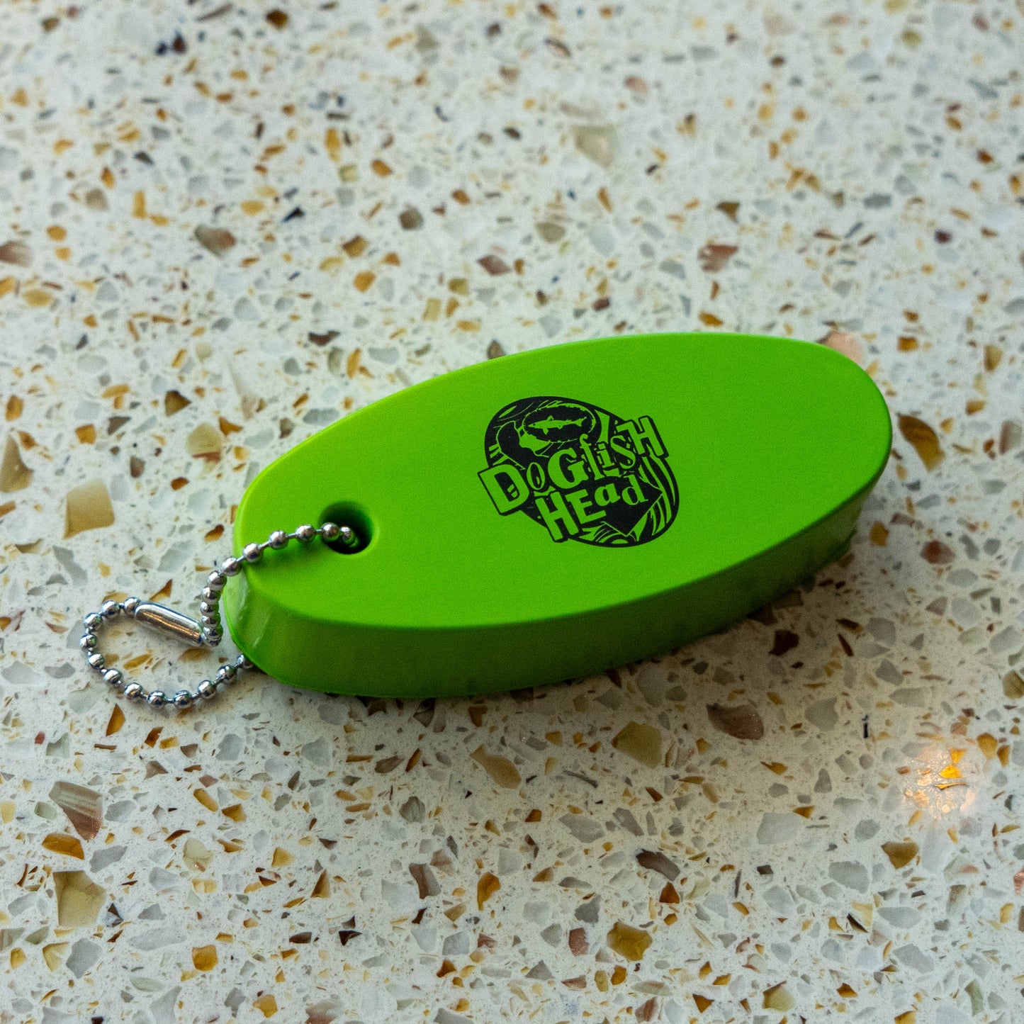 A bright lime green oval-shaped  keychain with a silver ball chain, featuring the 'Dogfish Head' logo, resting on a speckled terrazzo countertop.