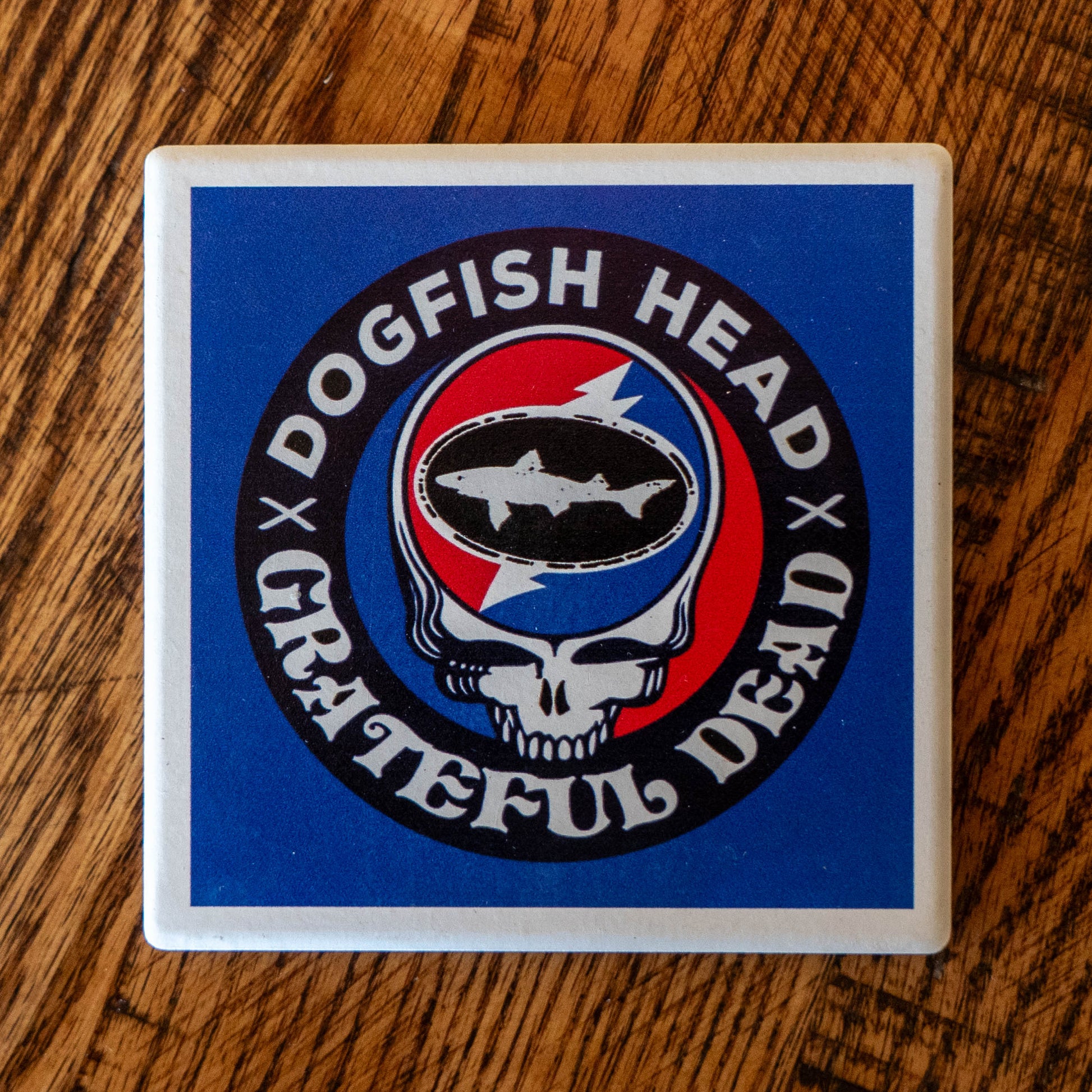 The Dogfish Head x Grateful Dead Ceramic Coaster by Paint the Town, Inc. features the iconic skull and dogfish logo with a red and blue lightning bolt inside an oval, surrounded by a blue border with white lettering on wood.