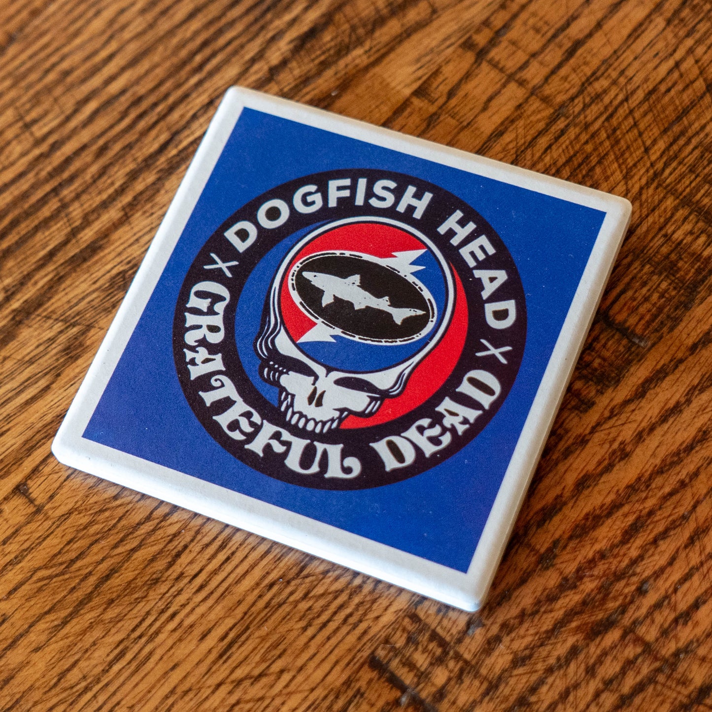The Dogfish Head x Grateful Dead Ceramic Coaster by Paint the Town, Inc. features a fish within a skull logo and "Dogfish Head Grateful Dead" text, resting stylishly on a wooden surface.