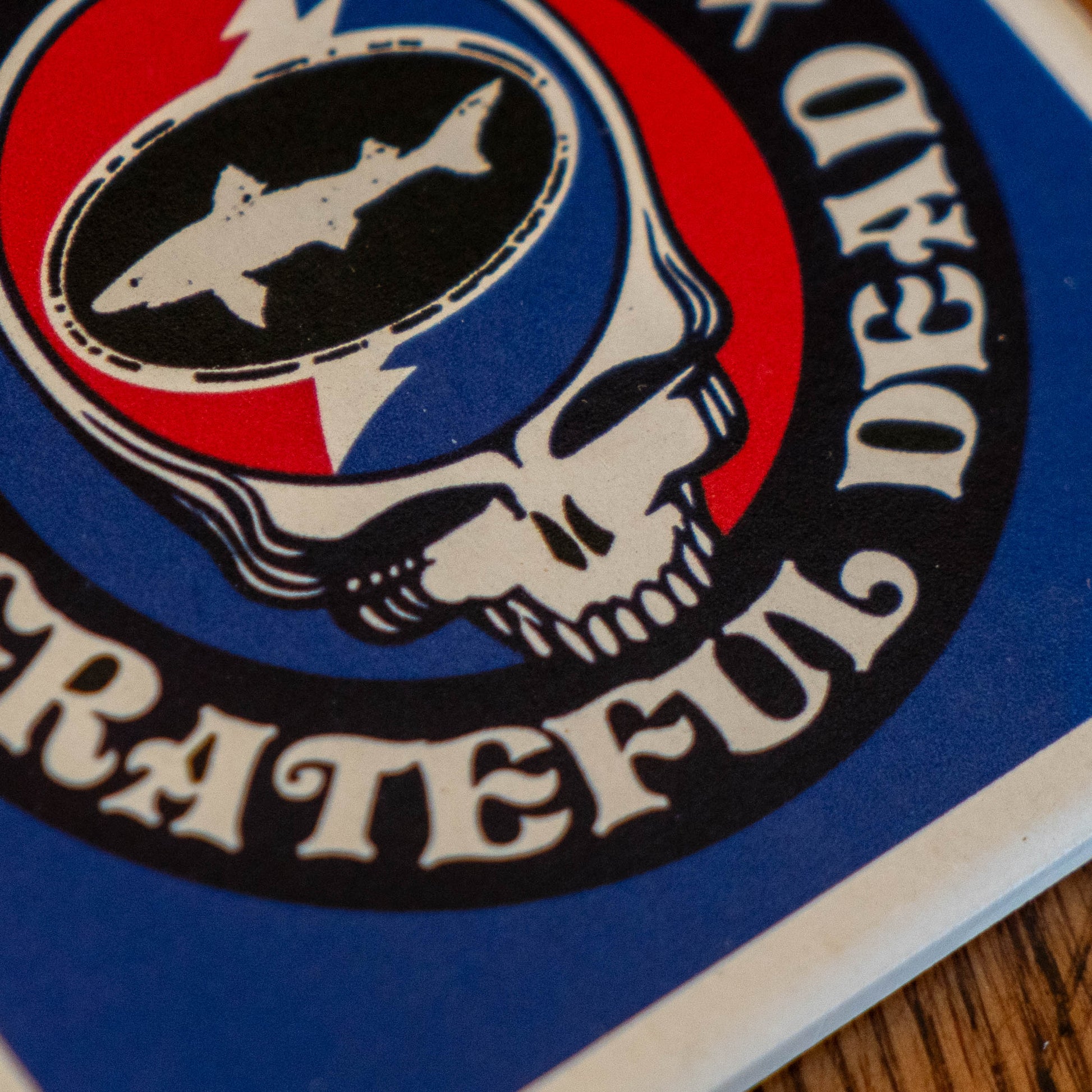 A close-up of the Dogfish Head x Grateful Dead Ceramic Coaster by Paint the Town, Inc., showing a skull with a lightning bolt, a shark silhouette inside the bolt, and the band's name in bold white letters on blue.
