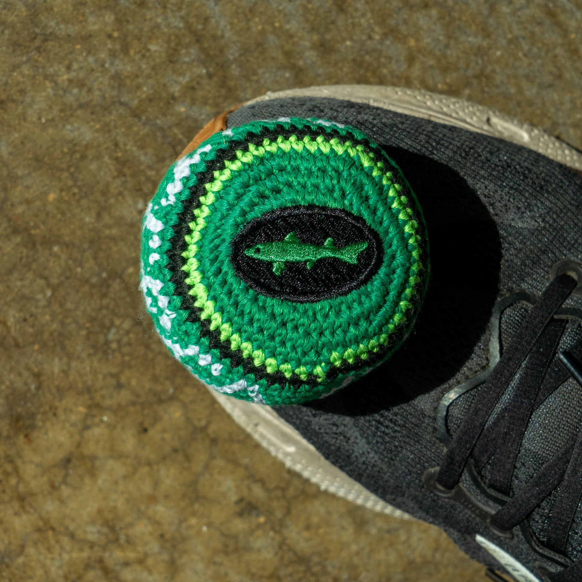 A close-up of a green crocheted ball on the heel of a black running shoe, placed on a textured brown stone surface. The cover features concentric circles in green, black, and white, with a green fish emblem in the center.