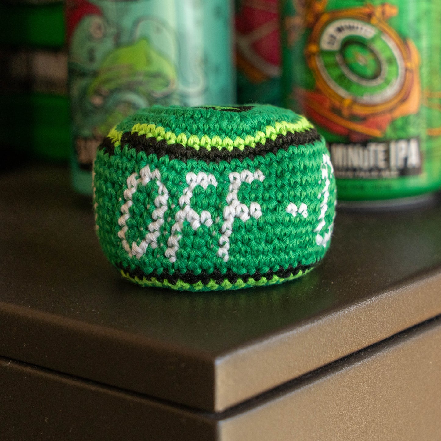 A green hand-knitted ball with white and black lettering that reads 'OFF-', sitting on a dark tabletop with blurred green beverage cans in the background.