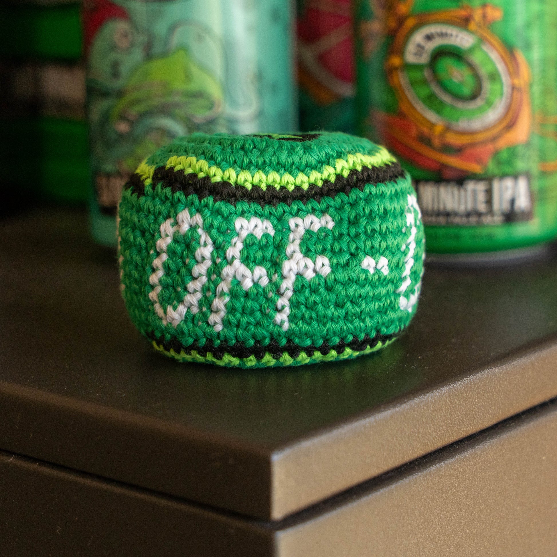 A green hand-knitted ball with white and black lettering that reads 'OFF-', sitting on a dark tabletop with blurred green beverage cans in the background.
