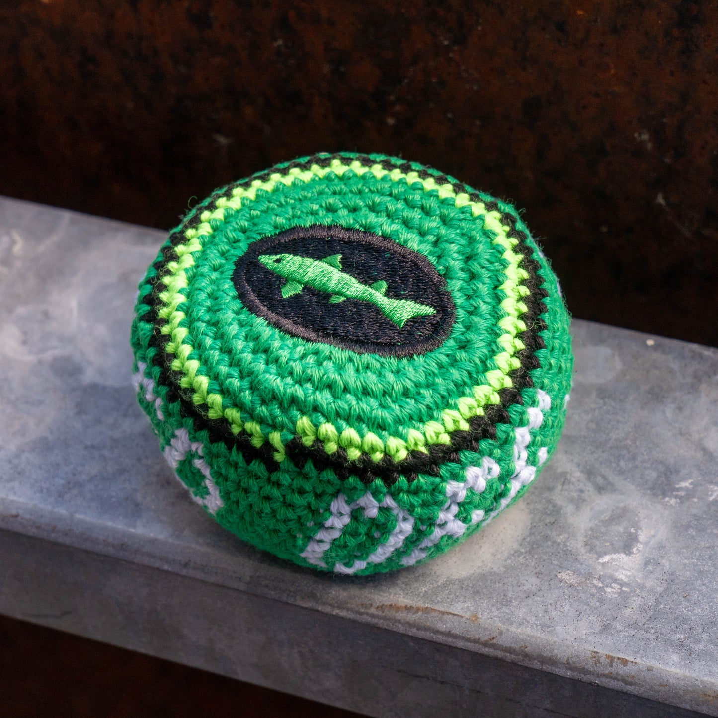 A green crocheted ball or with a black fish emblem on top, resting on a gray stone ledge against a dark brown textured background.