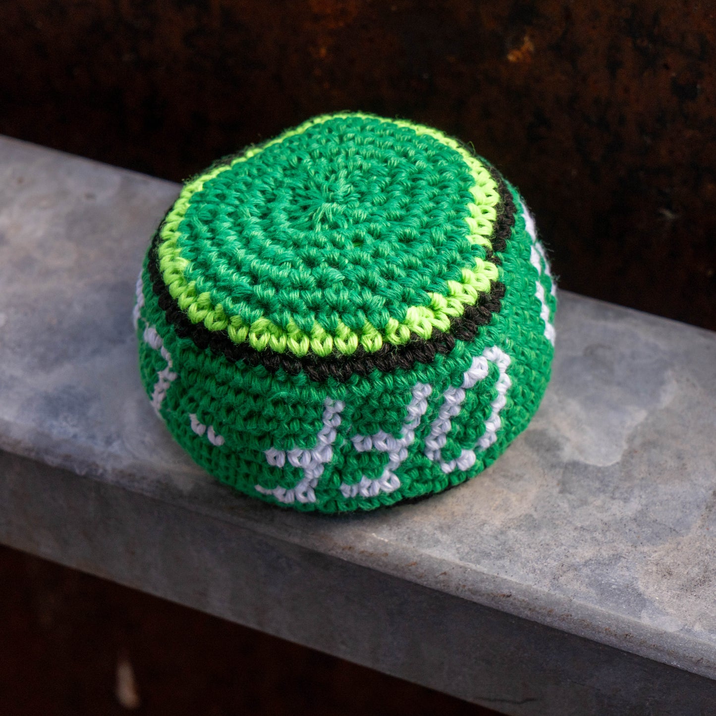 A handmade crocheted green ball with white and black lettering, resting on a gray stone ledge against a dark rusty background.