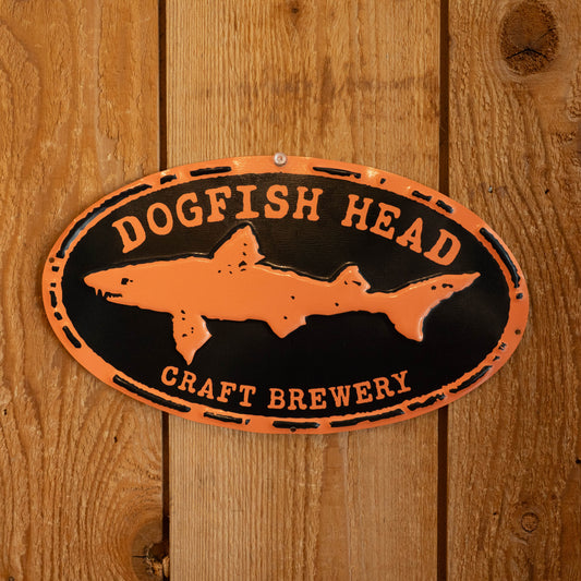 An oval metal sign for Dogfish Head Craft Brewery, featuring an orange shark silhouette on a black background with distressed orange lettering, mounted on a wooden plank wall.