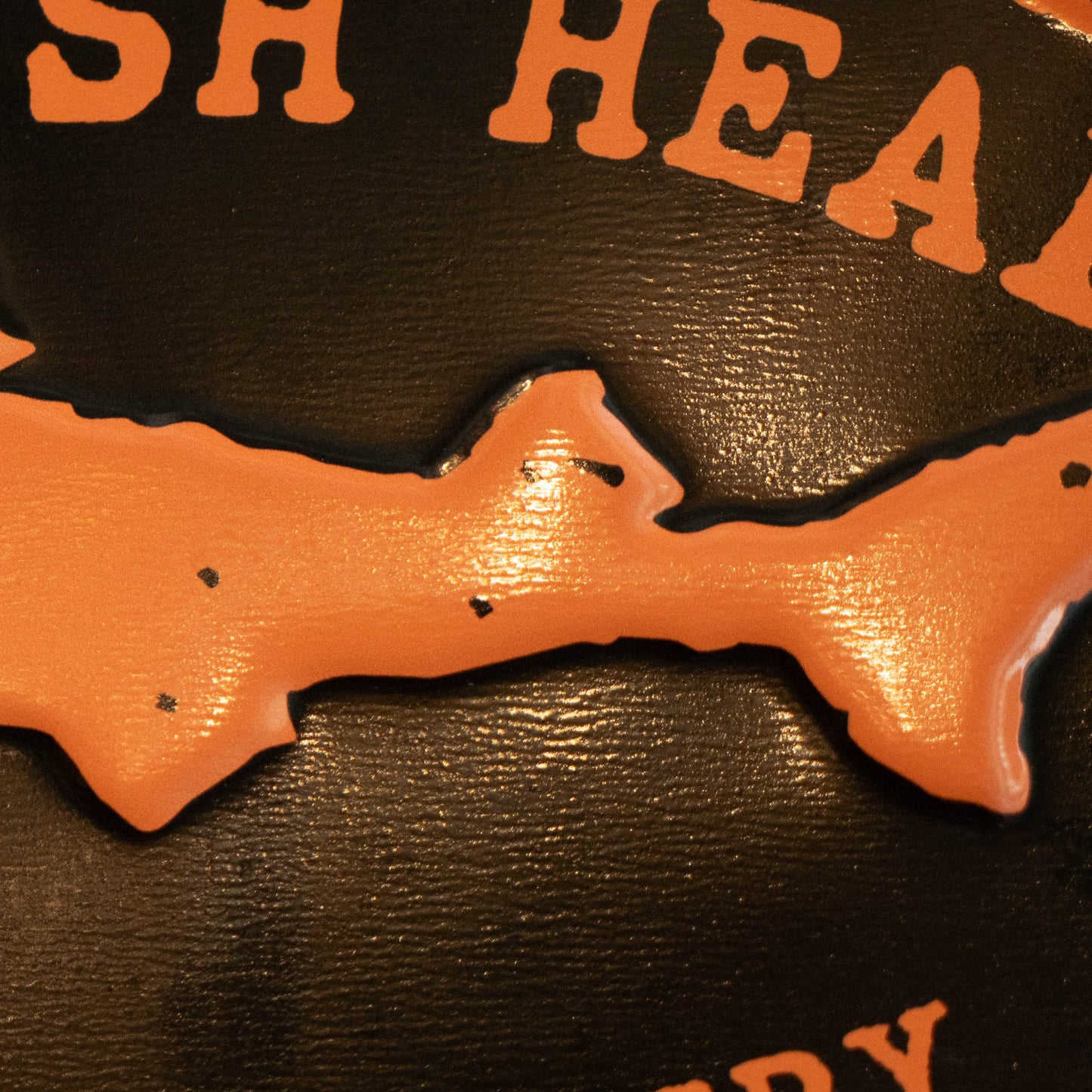 Close-up of a glossy orange fish-shaped plaque or emblem mounted on a dark textured background with partial orange lettering visible at the top and bottom.