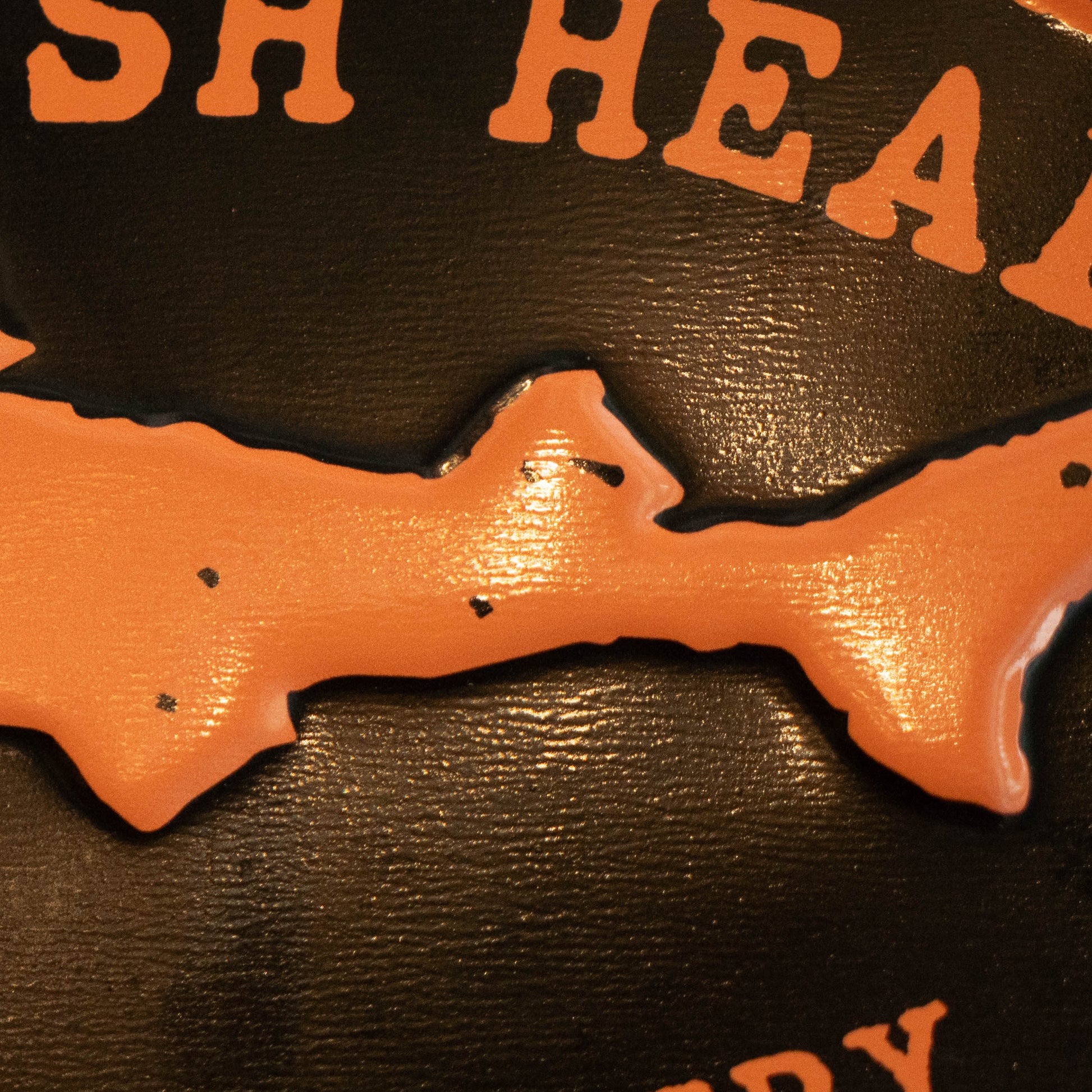 Close-up of a glossy orange fish-shaped plaque or emblem mounted on a dark textured background with partial orange lettering visible at the top and bottom.