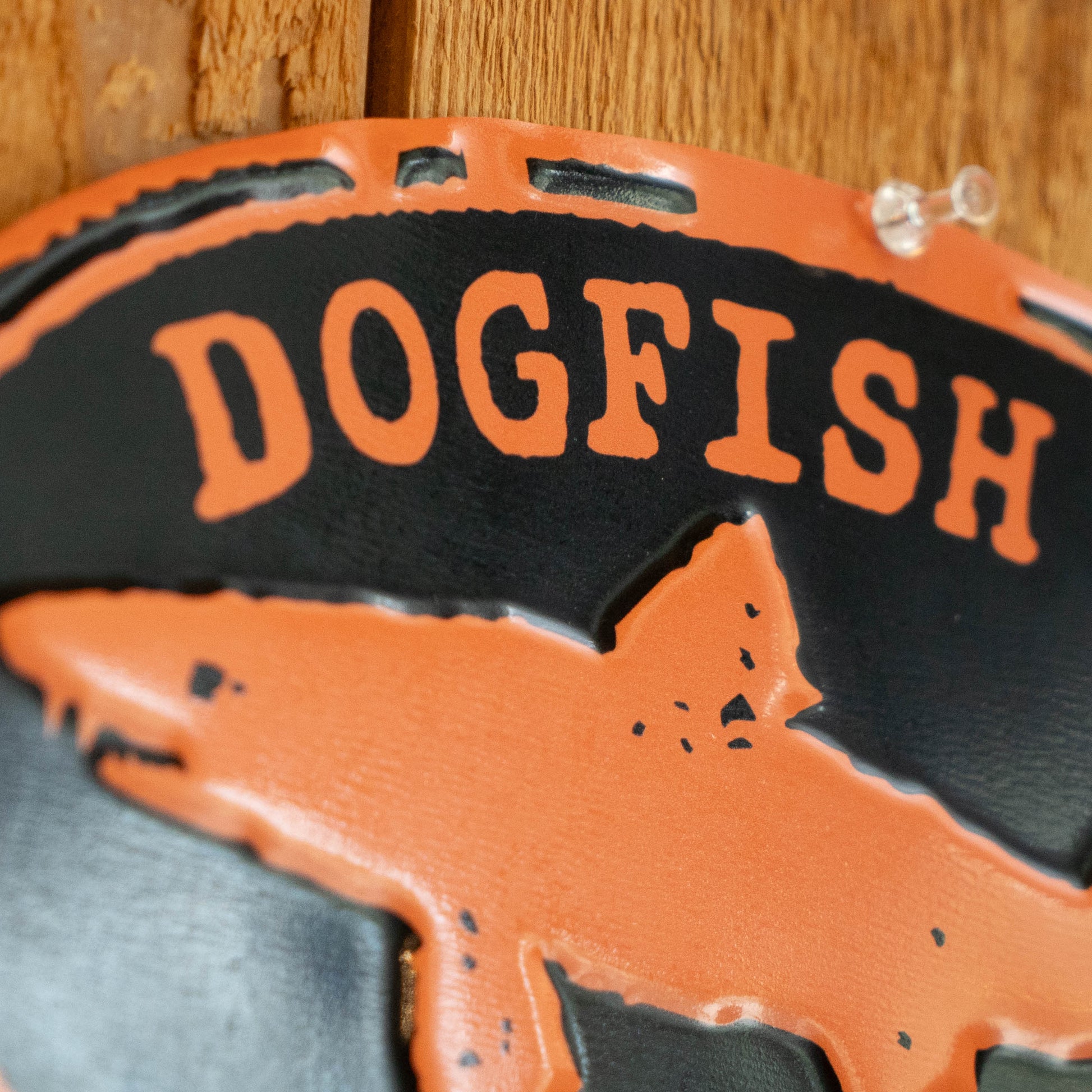 A close-up of a decorative metal sign featuring the words 'DOGFISH' in orange on a black background, with an orange dog silhouette and distressed cutout details, shown against a wooden surface.