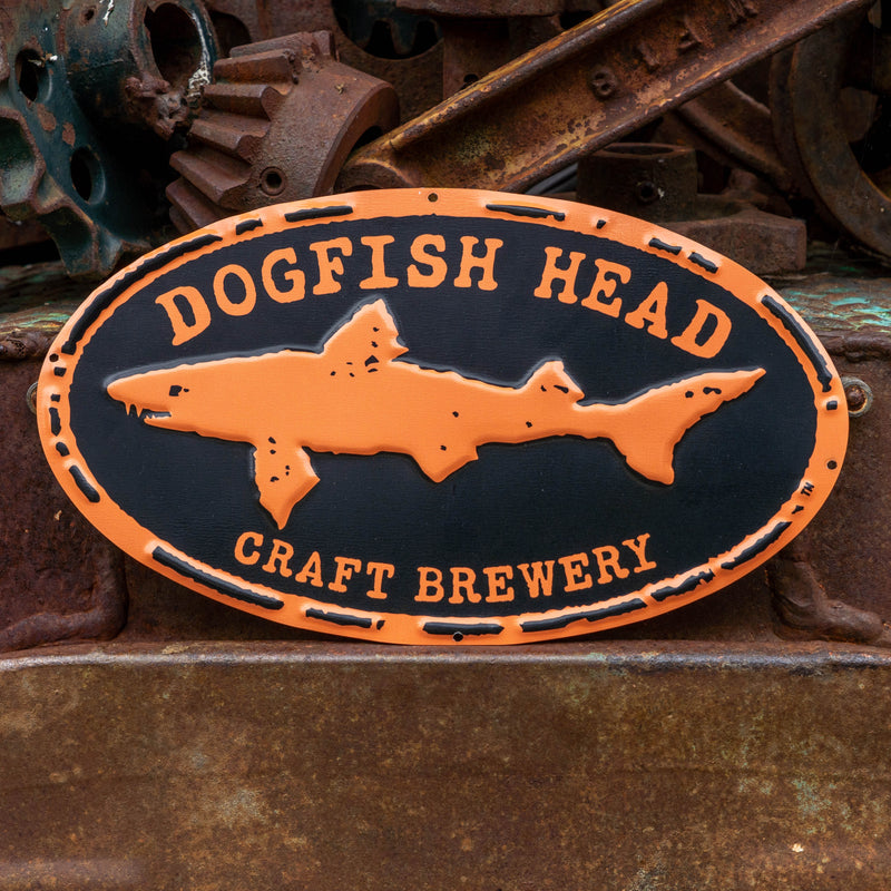 An oval metal sign for Dogfish Head Craft Brewery featuring an orange shark silhouette on a black background, photographed against a rustic rusty metal surface.
