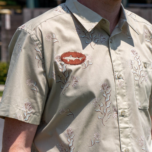 Someone is wearing a Patagonia M Brown Go To Shirt—an organic cotton, light beige short-sleeve button-up with a floral pattern and a brown oval patch featuring a white fish on the chest. The photo is taken outdoors.