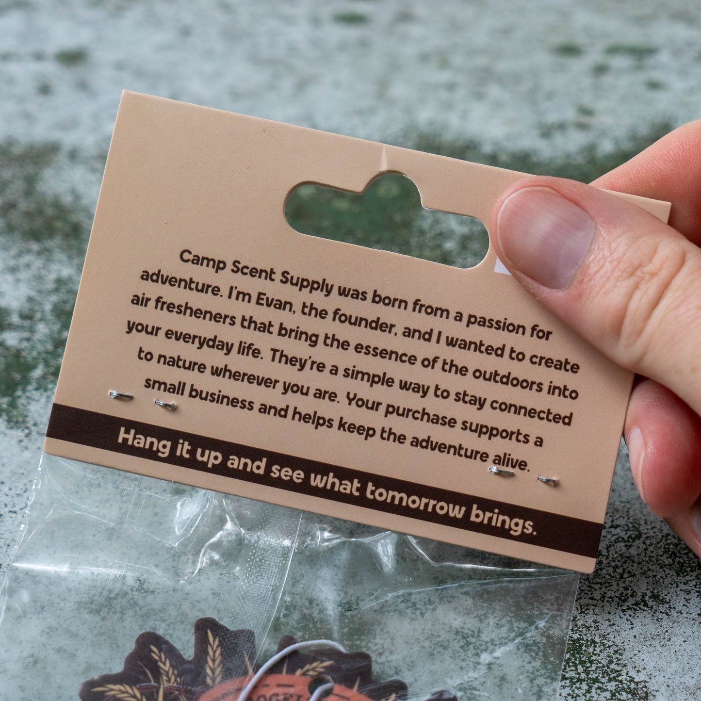 A hand holding a kraft paper hanger card with printed company text and two small pins attached, above a clear plastic bag containing an orange and brown patch. The background is a textured gray stone surface.