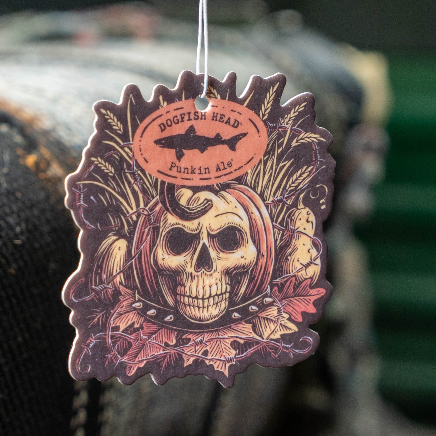 A die-cut hanging air freshener tag featuring a skull, corn, fish, and autumn leaves design with the text 'Dogfish Head Punkin Ale' on a circular label, shown against a blurred outdoor background.