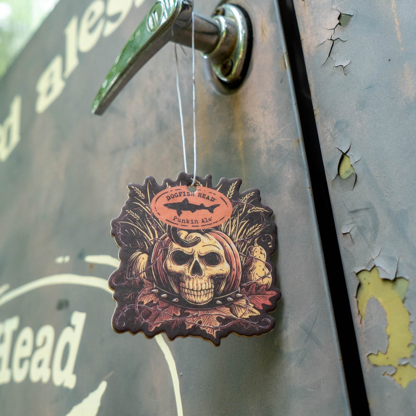 A decorative wooden air freshener shaped like a skull wearing a bandana hangs from a metal latch on a rustic metal door. Features a detailed skull illustration with a circular logo at the top that reads 'Dogfish Head Punkin Ale'.