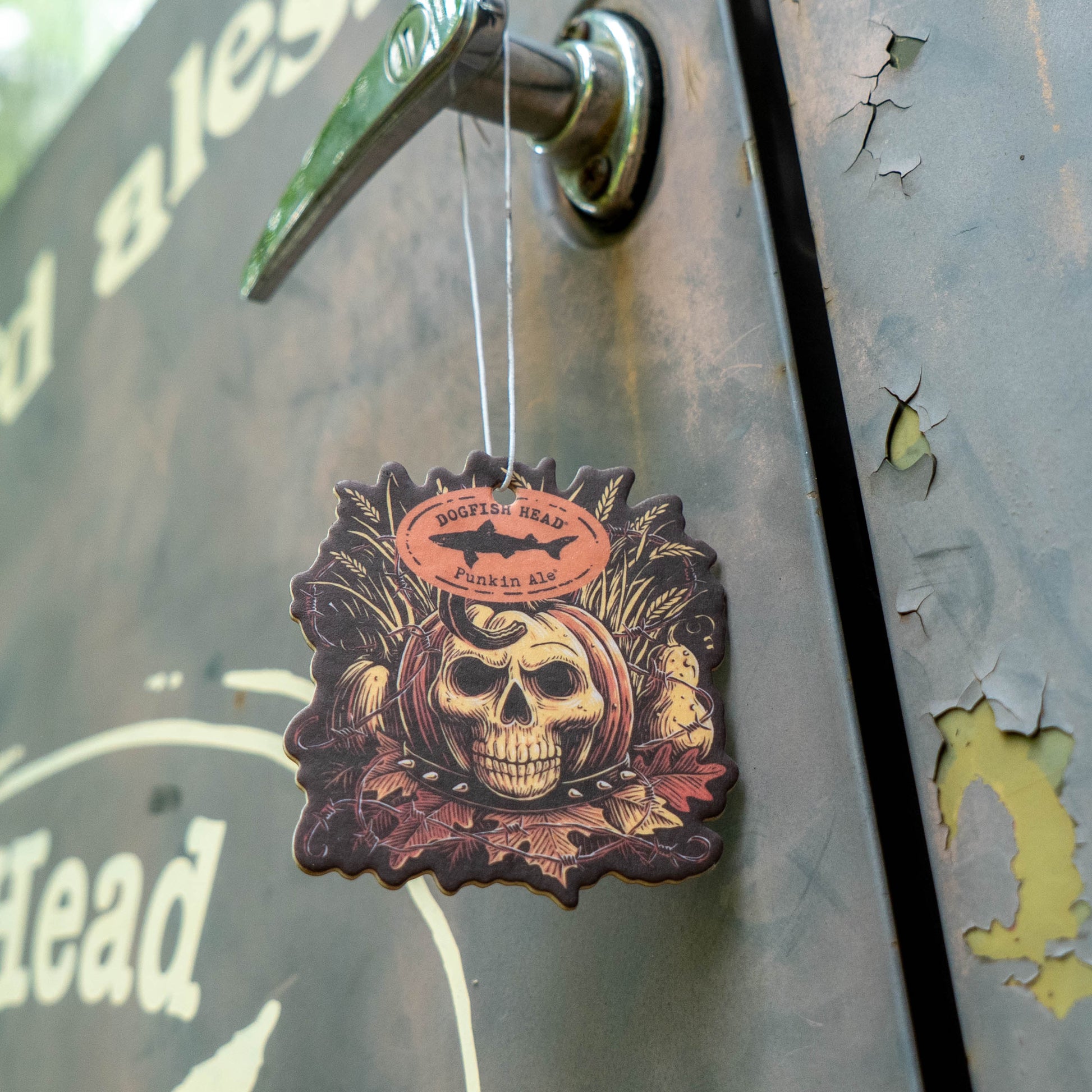 A decorative wooden air freshener shaped like a skull wearing a bandana hangs from a metal latch on a rustic metal door. Features a detailed skull illustration with a circular logo at the top that reads 'Dogfish Head Punkin Ale'.