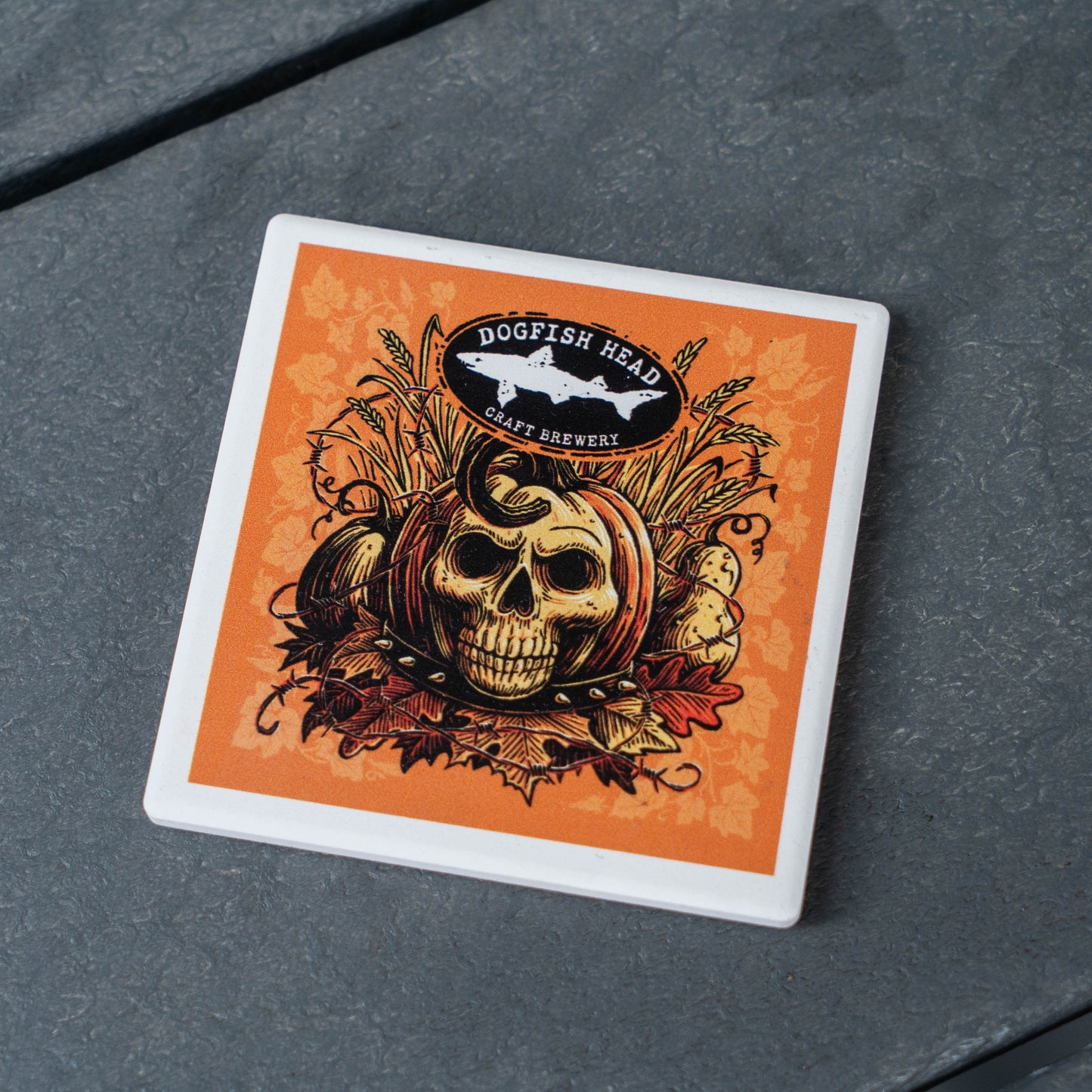 A square white coaster with rounded corners featuring an orange and black graphic design of a skull, autumn leaves, and a shark logo that reads 'Dogfish Head Craft Brewery', placed on a dark gray textured surface.