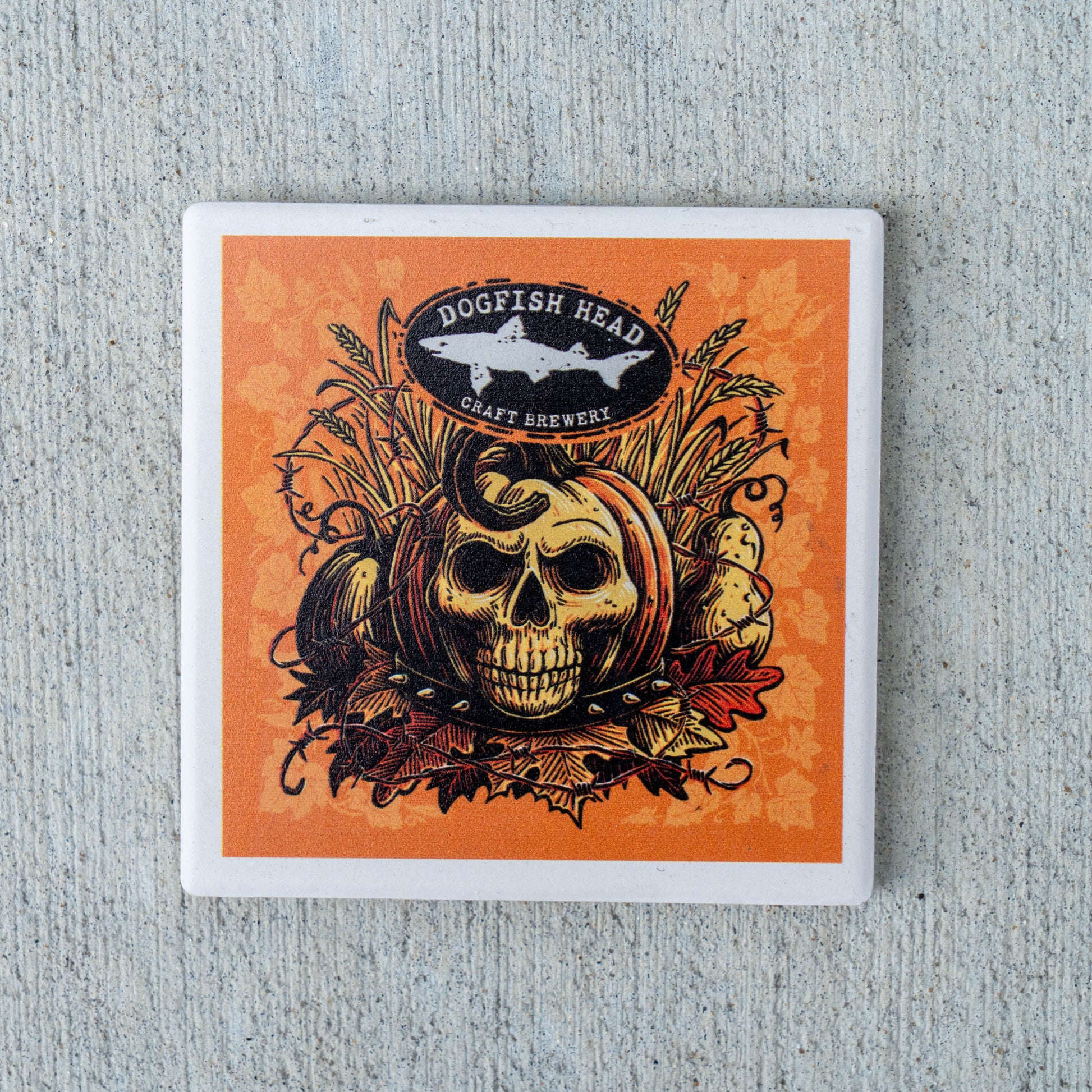 A square coaster with a white border featuring an orange and black graphic design of a skull in a pumpkin, wheat, and autumn leaves, with the Dogfish Head Craft Brewery logo at the top, shown on a textured gray background.