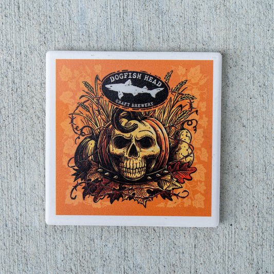 A square coaster with a white border featuring an orange and black graphic design of a skull in a pumpkin, wheat, and autumn leaves, with the Dogfish Head Craft Brewery logo at the top, shown on a textured gray background.